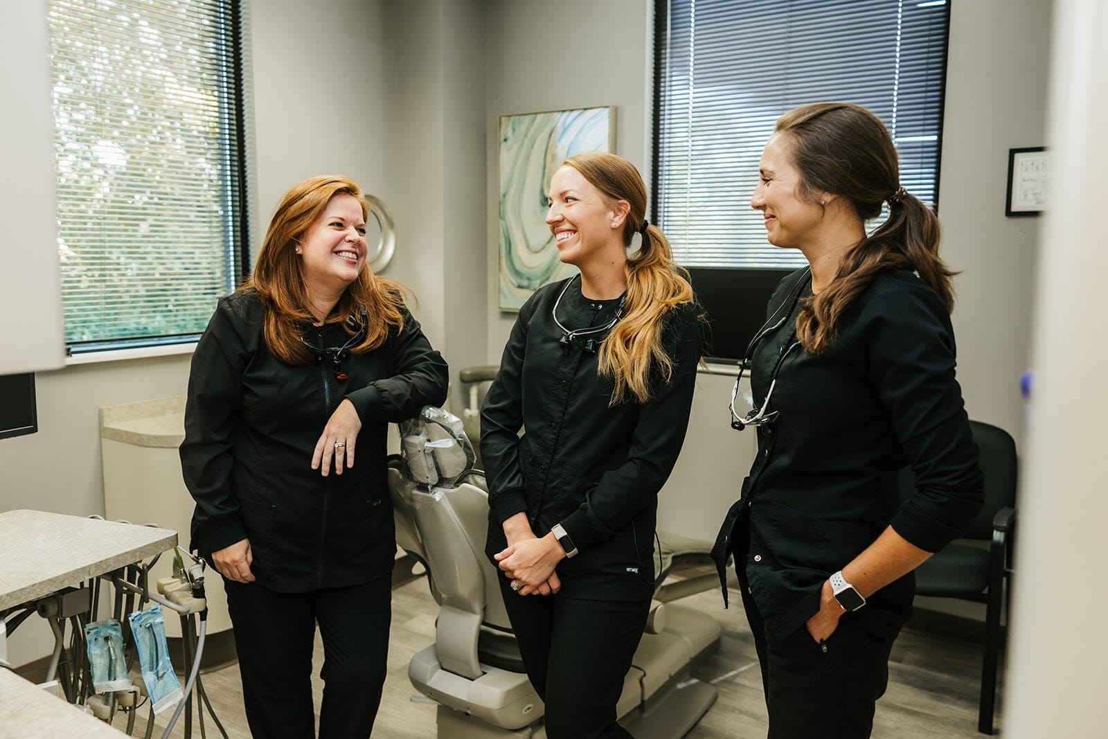 Three members of a dental office team standing together in a treatment room, smiling and talking.