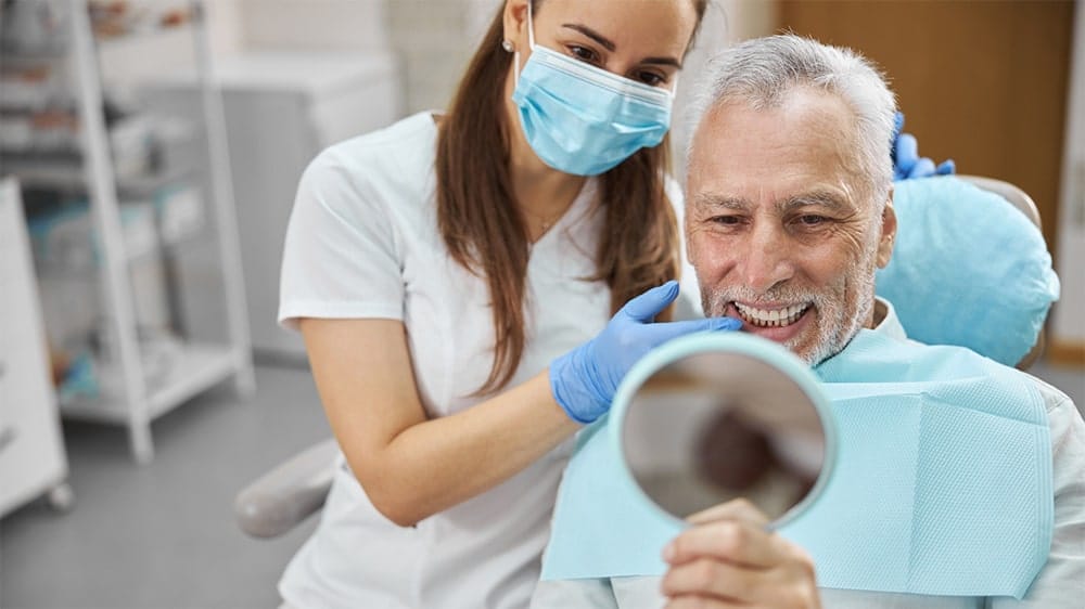 A senior male patient smiling at his reflection in a mirror while a dental professional assists