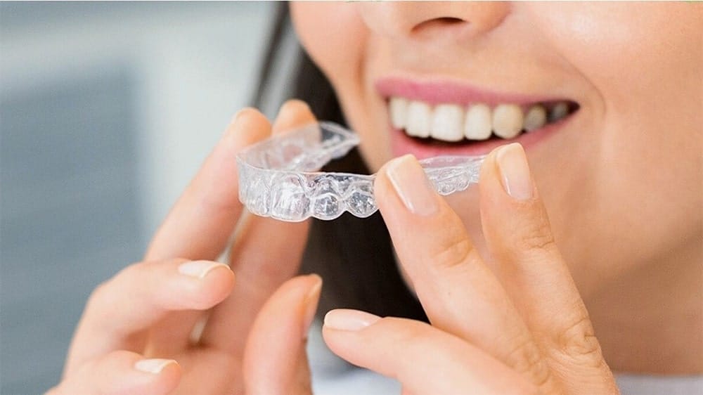 A woman smiling while holding a clear aligner near her mouth