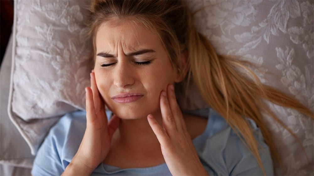 Woman lying in bed holding her jaw in pain with a pained expression