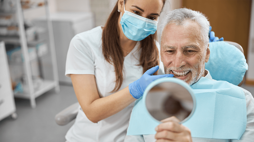 A senior male patient smiling at his reflection in a mirror while a dental professional assists