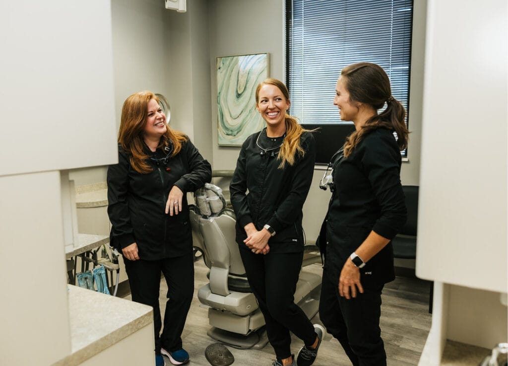 Three female dental professionals in black scrubs smiling and talking inside a dental exam room